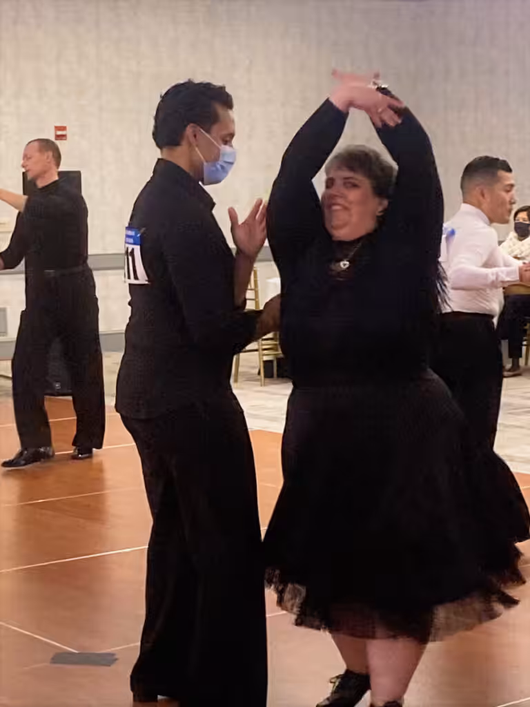 A dance couple dressed in black at an indoor event with other dancers in the background.