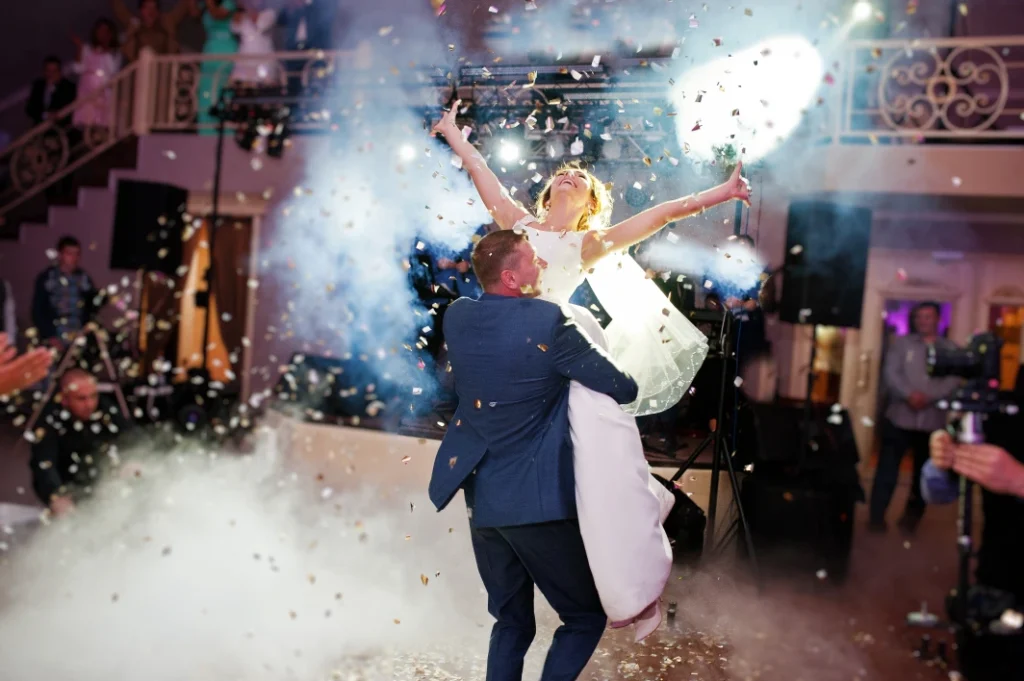 Groom lifts the bride in a white dress during their wedding dance. Confetti fall around them with smoke effects in the background.