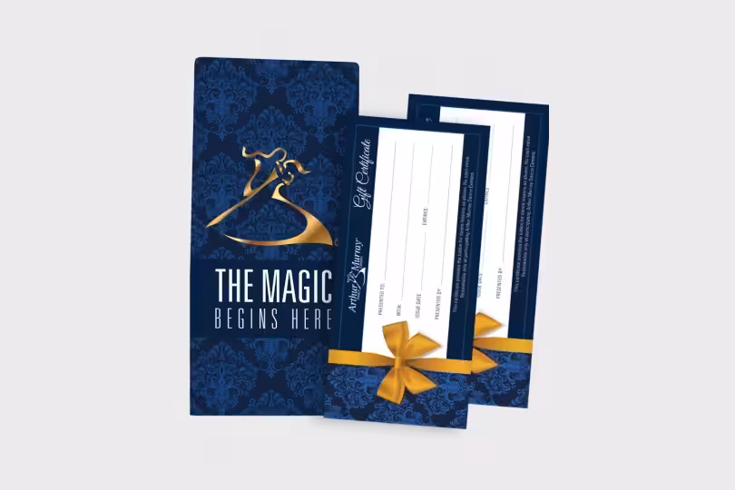 Two gift certificates with gold bows rest against a blue and gold patterned envelope that reads The Magic Begins Here.