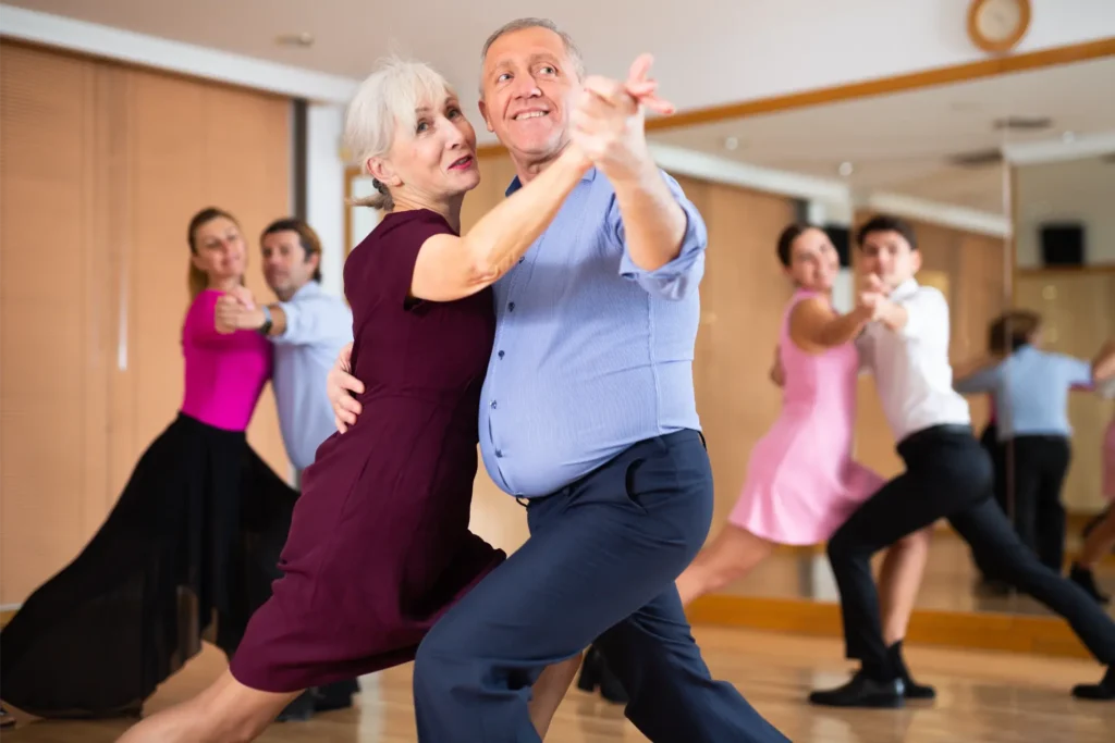 Older couple dancing in front of a mirror with other couples practicing ballroom dance in the background of a studio.