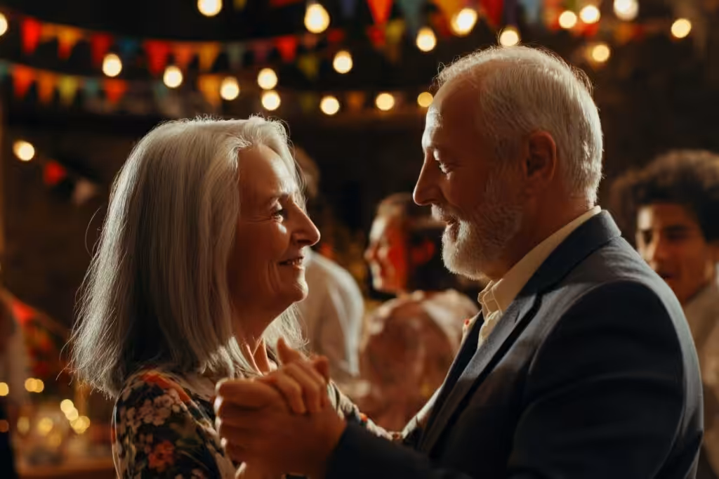 An older couple dances, looking at each other. He wears a dark suit, she a floral top. Lights and banners hang.