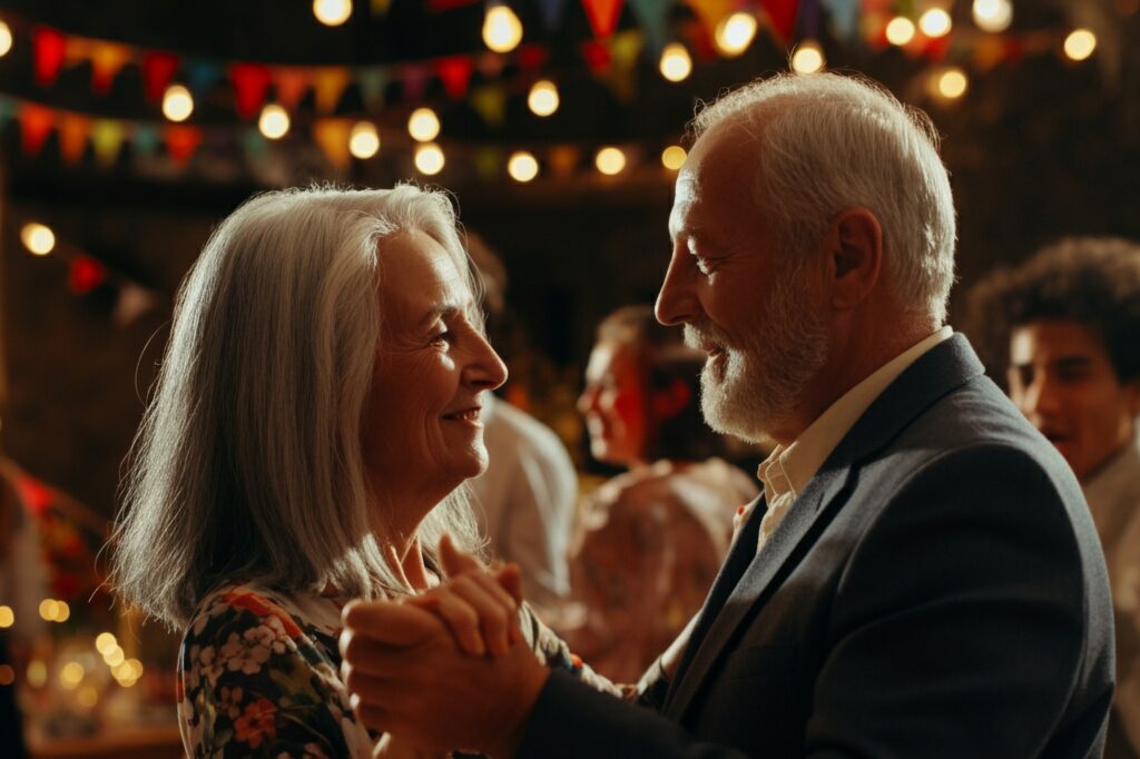 An older couple dances, looking at each other. He wears a dark suit, she a floral top. Lights and banners hang.