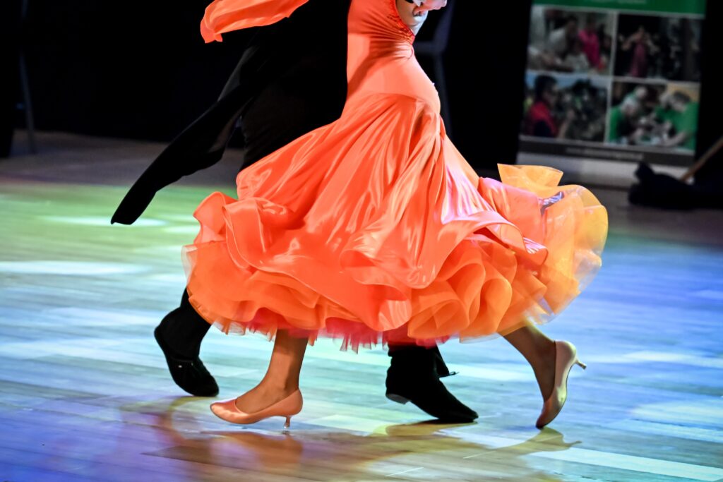 A couple ballroom dances. The woman wears a bright orange dress and high heels. The man wears black pants.