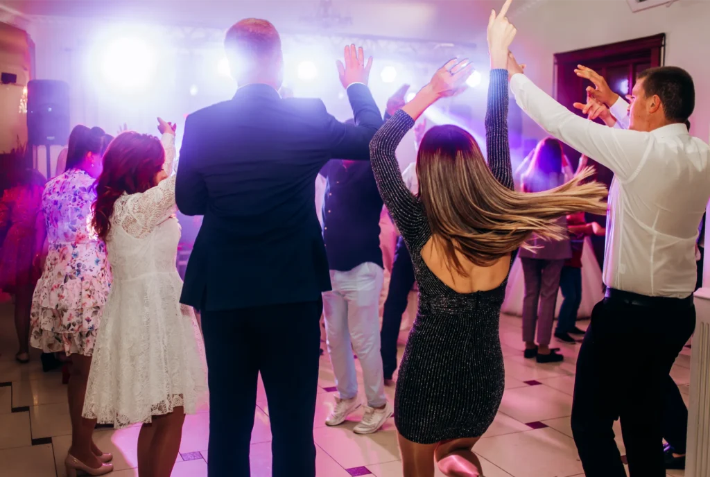 A diverse group of men and women dance with their hands raised under bright lights at a party.