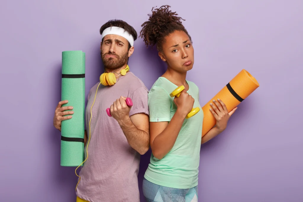 A bored man and woman in workout clothes hold dumbbells and yoga mats, looking annoyed.