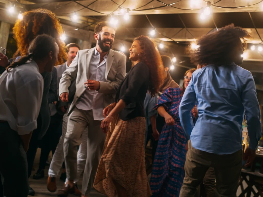 A lively outdoor dance party at night with a group of friends laughing and dancing under string lights.