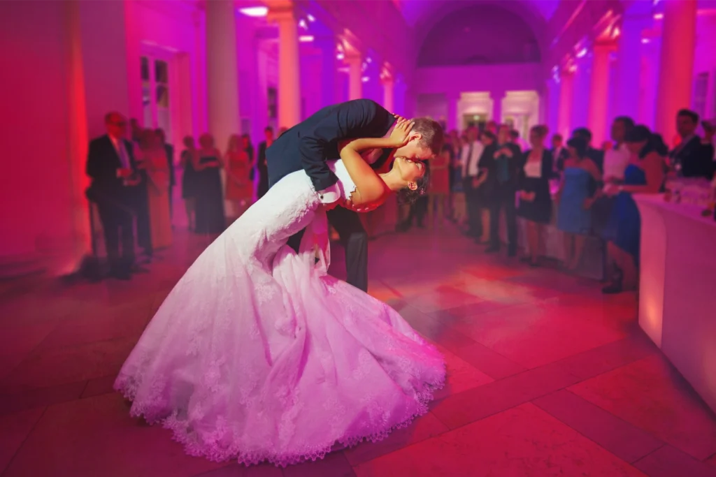 A couple in formal wedding attire performs a deep dance dip on a pink-lit ballroom floor surrounded by guests.