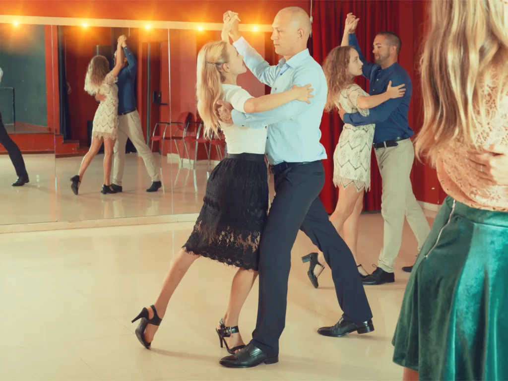 Couples practicing ballroom steps in a mirrored studio: partners holding dance frame positions and moving across the floor.