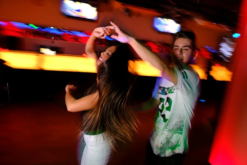 Two young people, a woman with long dark hair and a man in a white t-shirt, dancing energetically in a club with red lighting.