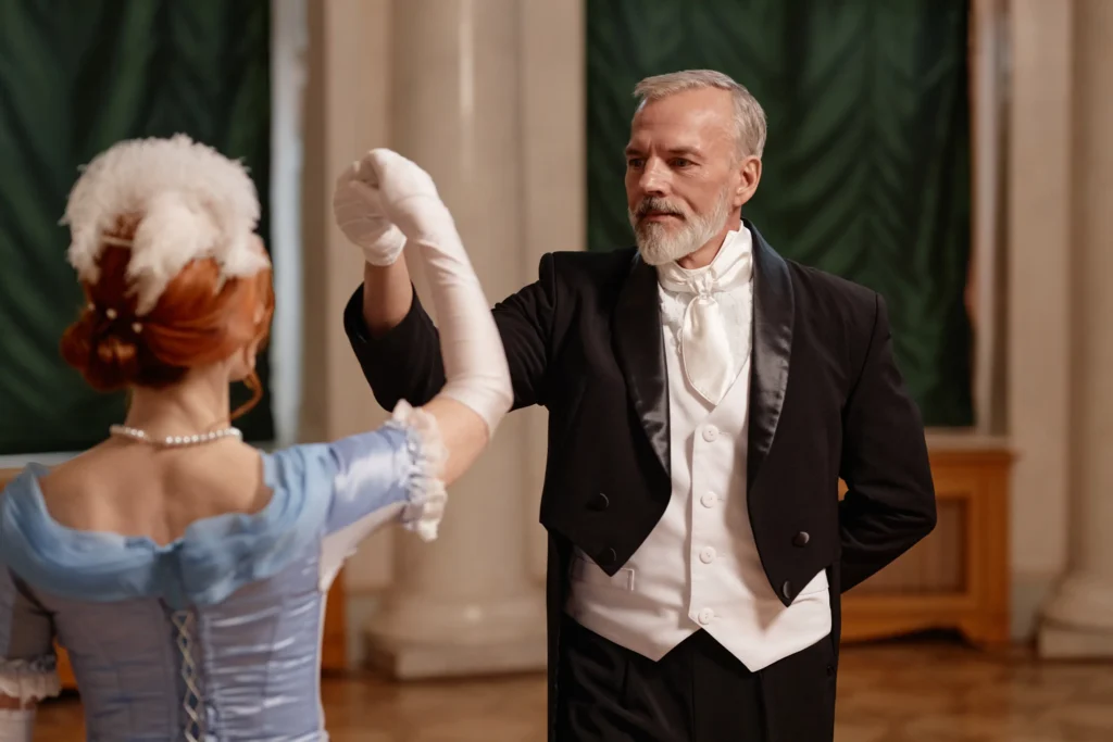 A mature man in a black tuxedo and white cravat dancing with a woman in a light blue historical gown and feathered hairpiece.
