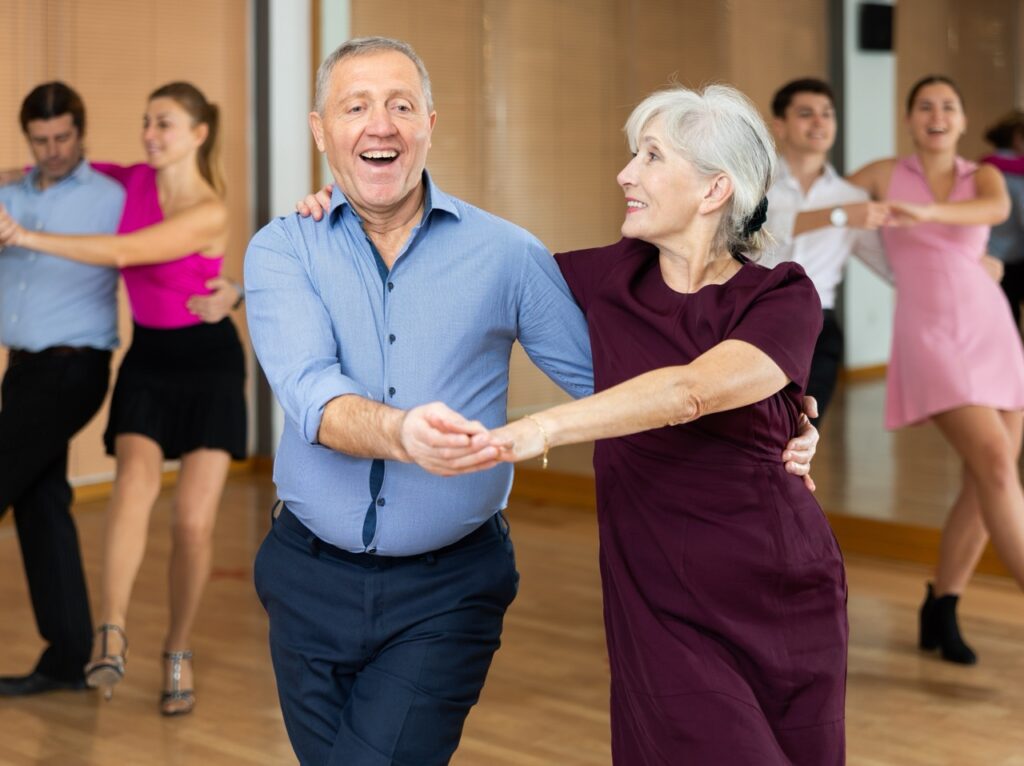 Smiling older couple ballroom dancing together in a studio while other students practice in the background.