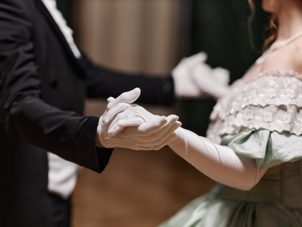 Close-up of a couple in formal attire holding hands in a ballroom dance position, both wearing white gloves.